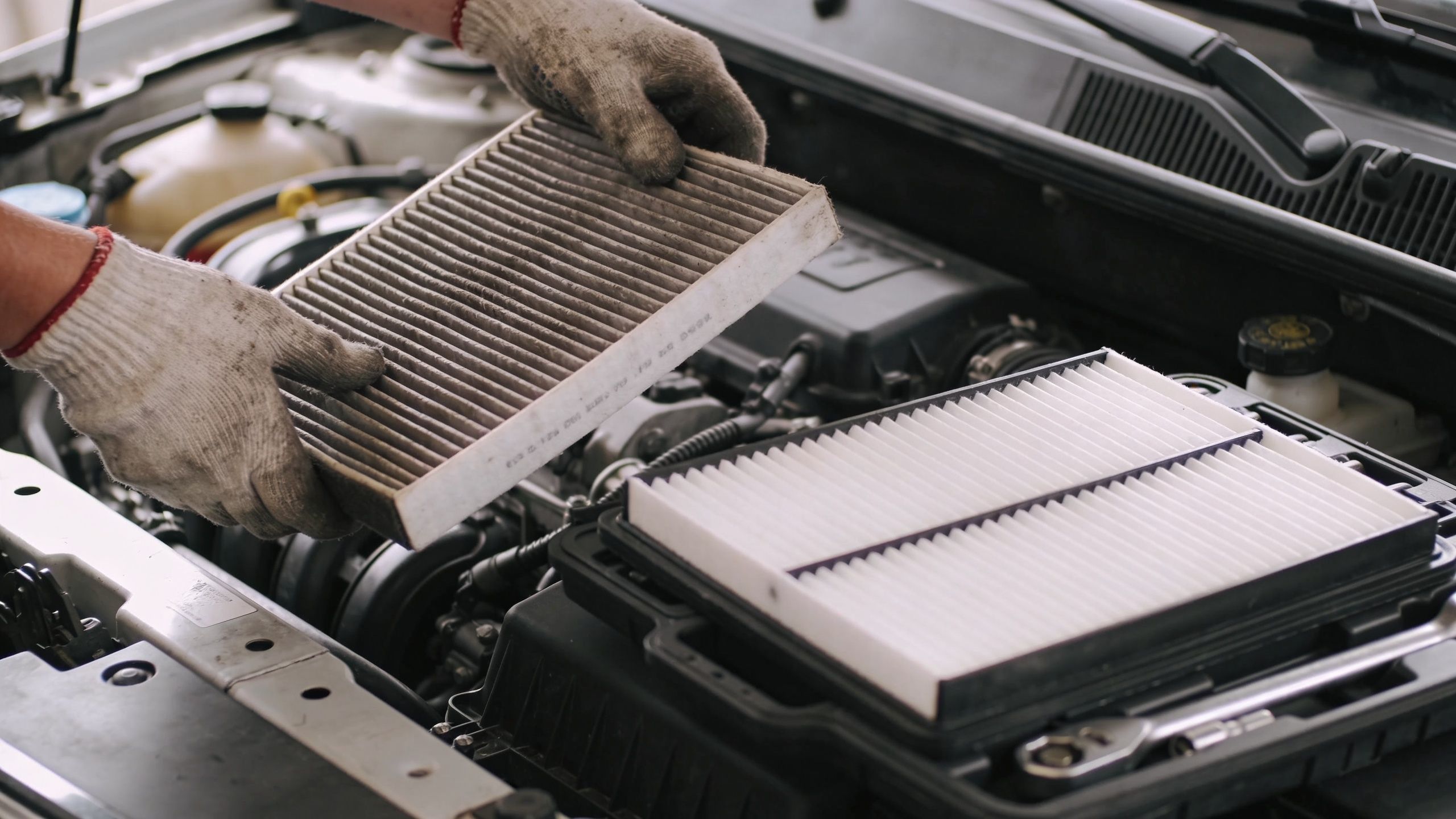 A mechanic wearing work gloves holds a dirty used car engine air filter above a clean new one.