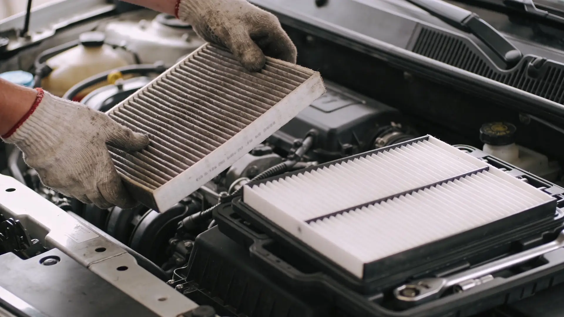 A mechanic wearing work gloves holds a dirty used car engine air filter above a clean new one.