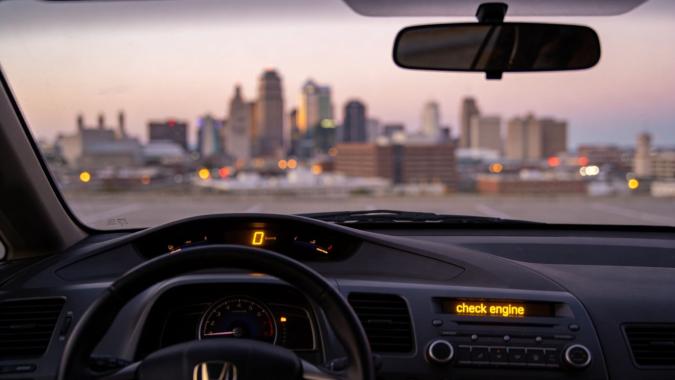 A view from inside a car parked facing a city skyline during sunset with a check engine warning