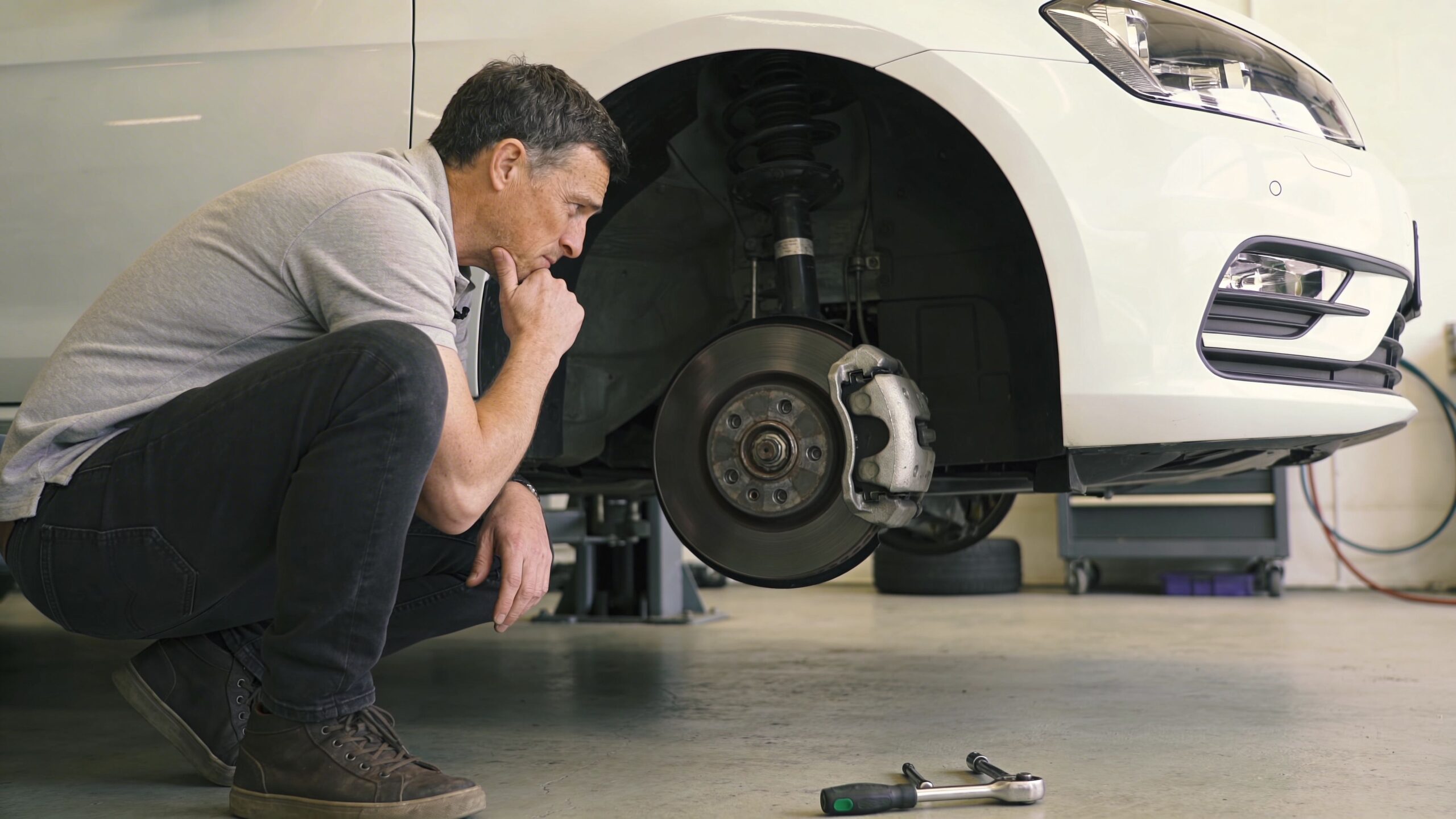 A professional mechanic crouches and examines the brake system of a lifted car in an auto shop.