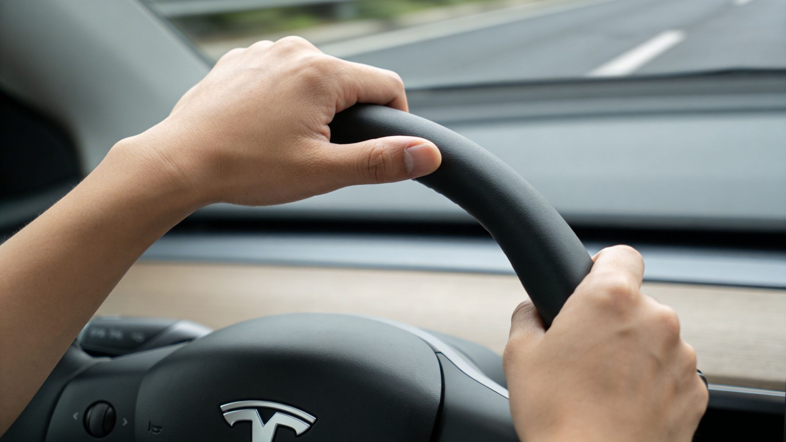 A driver's hands gripping the steering wheel of a Tesla car while driving on a road.